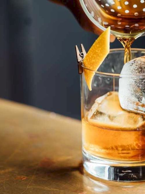 A cocktail being poured into a glass with ice and a citrus twist garnish, set on a bar counter.