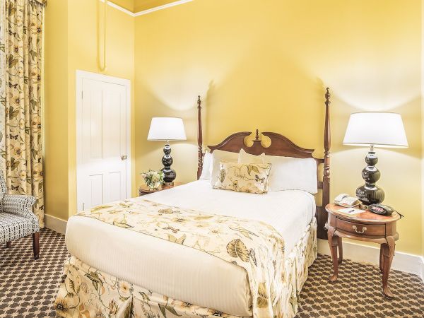 A cozy room with yellow walls, a bed with floral bedding, two lamps, a chair, a window with patterned curtains, and a decorative carpet.