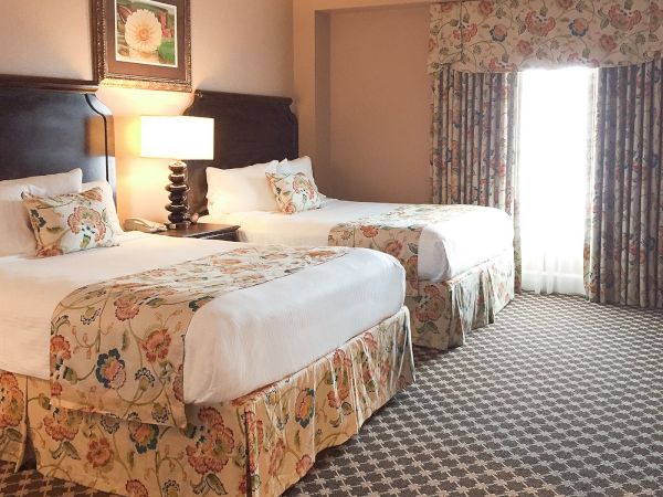 The image shows a hotel room with two beds, floral bed covers, patterned carpet, a nightstand with a lamp, and large curtained window.