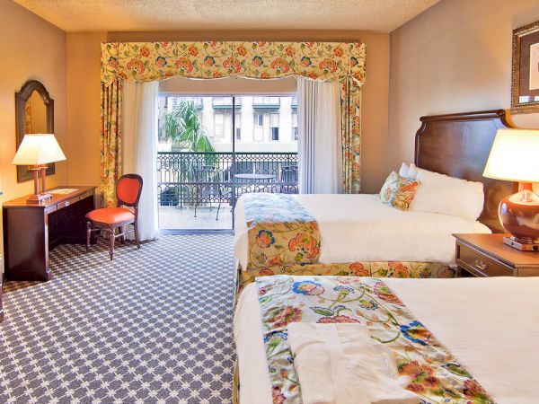 A hotel room with two beds, floral decor, a TV, desk, lamps, and a balcony with a view outside.