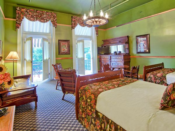 A green-themed room with floral decor, two beds, a chandelier, wooden furniture, and a patterned carpet, featuring open doors to a balcony.