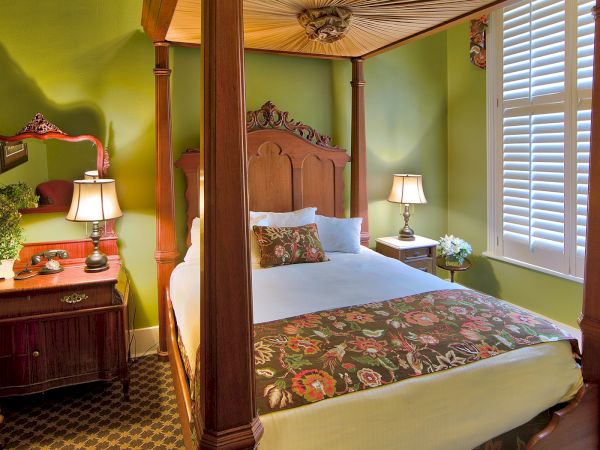 A cozy bedroom with a four-poster bed featuring floral bedding, green walls, a dresser with a mirror, and lamps on nightstands near shutters.