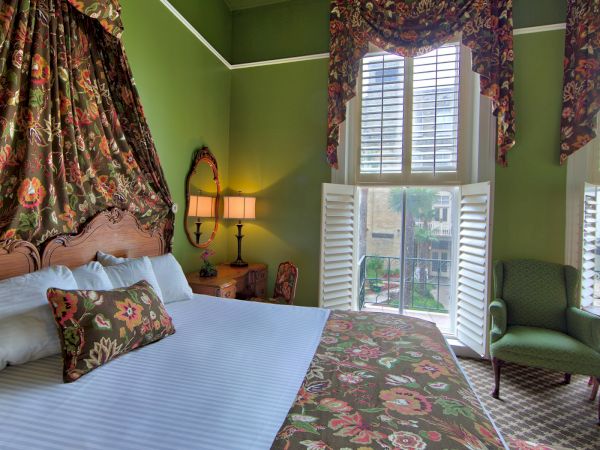 A cozy bedroom with green walls, floral decor, large windows with shutters, an armchair, a bed with white linens, and a floral bedspread.