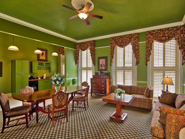 A living room with green walls, a dining table, couches, a ceiling fan, and floral curtains in a classic style.