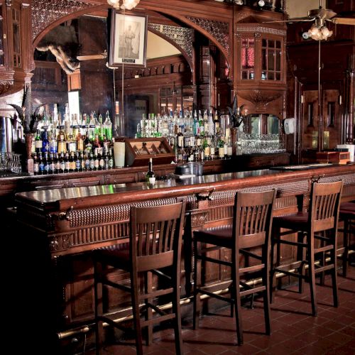 A dim, classic bar with wooden stools, glassware, and bottles lining the back wall; warm, inviting tavern ambiance.