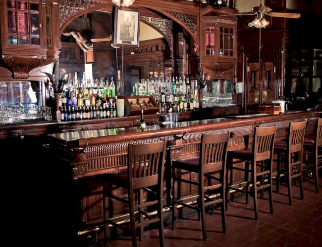 A dim, classic bar with wooden stools, glassware, and bottles lining the back wall; warm, inviting tavern ambiance.