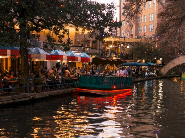 People dining along a canal at dusk; a boat glides by under a lit bridge, restaurant tents line the water with warm, inviting glow.