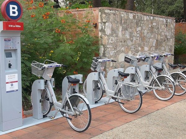 A row of gray bicycles docked at a station with a nearby blue and red charging/payment kiosk, set against a stone wall and greenery.