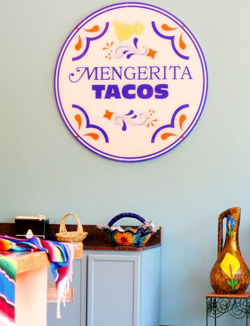 Colorful cafe interior with a large circular sign &ldquo;Mengerita Tacos,&rdquo; menu boards, and vibrant textiles on a light blue wall. End.