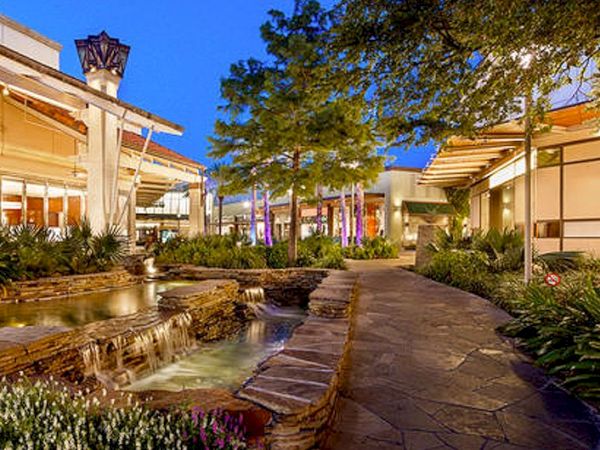 A sunny outdoor shopping courtyard with shops, a stone path, lush plants, and a small water feature along a walkway at dusk.