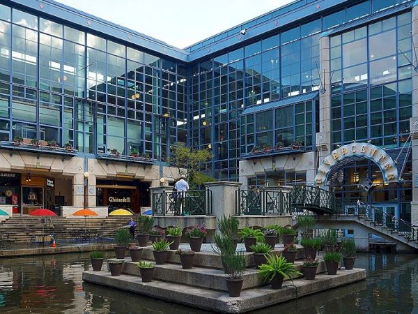A modern glass-building courtyard with water features and potted plants arranged on tiered platforms, plus a curved metal entrance and small waterfalls, stylish and serene.