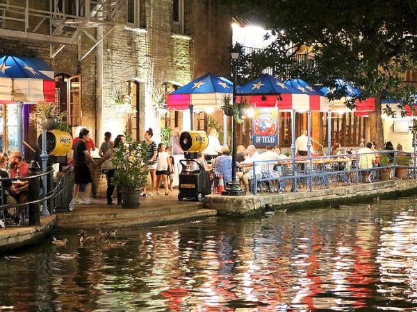People dining at riverside cafes under colorful umbrellas at night, reflections dancing on the water. A lively waterfront scene.