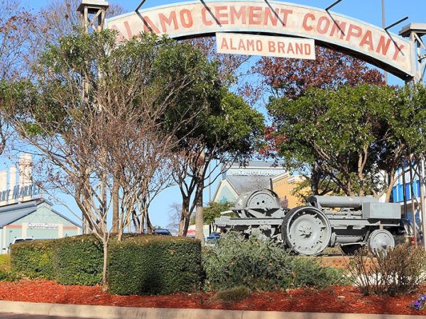 Alamo Cement Company entrance sign arch over a landscaped area with trees, a small vintage engine statue, and factory buildings in the background.
