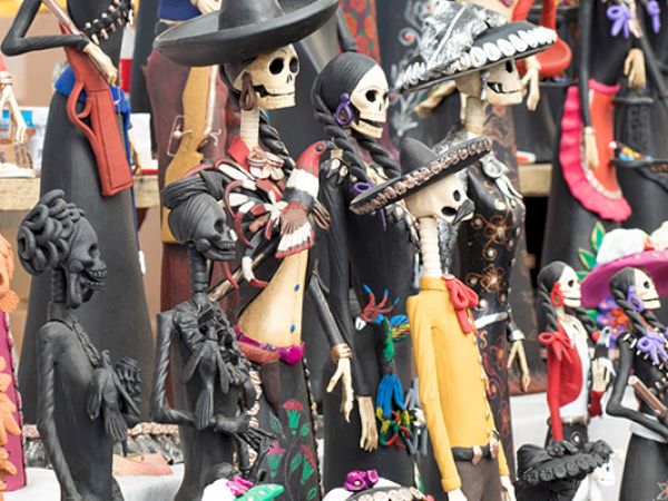 A display of colorful handmade figurines, including skeletal figures in hats and cloaks, plus vibrant magenta, yellow, and orange outfits, all in a whimsical, spooky market scene.