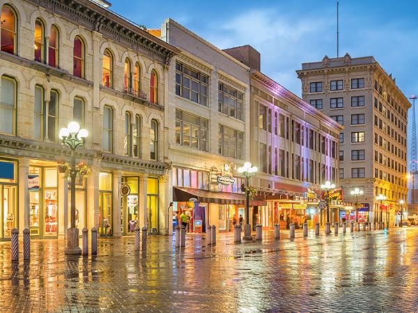 A lively street scene at dusk with historic buildings, storefronts, and colorful lights reflecting on a wet pavement, bustling with pedestrians.