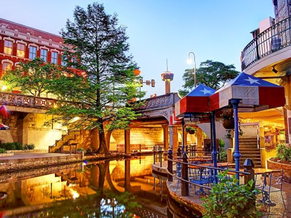 A charming outdoor plaza with a canal, trees, lamps, and colorful umbrellas over seating, reflecting warm lights on the water.