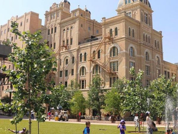 A grand beige brick building with arched windows sits behind a green park with trees, people walking, and a fountain nearby, sunny day.