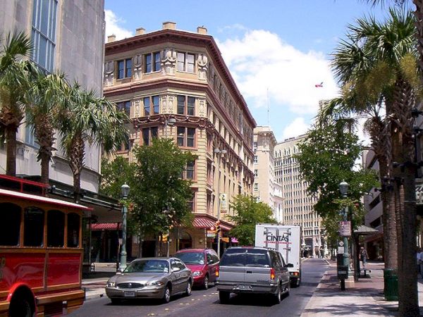 A sunny street scene with palm trees, a red trolley, and a mix of historic and modern buildings, crowded with cars and pedestrians.