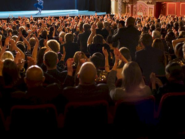 A large audience in a theater or concert hall, many hands raised, facing a stage with a performer in the spotlight.