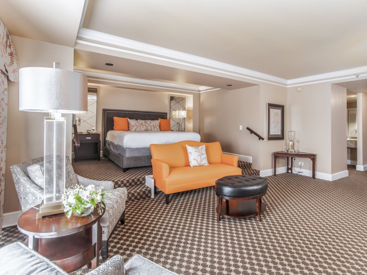 A luxury hotel suite with a king bed, orange sofa, matching ottoman, patterned carpet, and stylish seating area in warm tones.