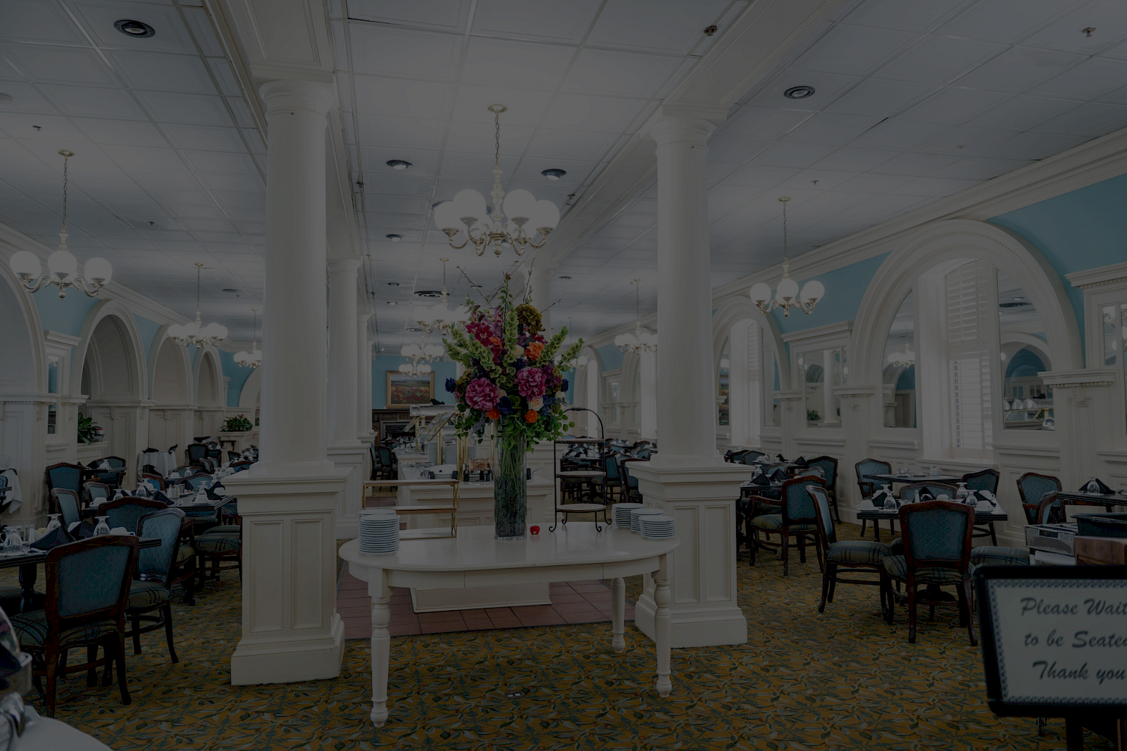 A bright, elegant dining hall with white columns, chandeliers, arched windows, blue accents, and a floral centerpiece on a white table.