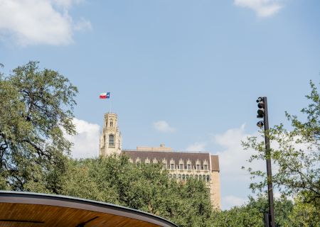 A tall, historic building with a flag on top peeks through green trees under a blue sky with a few clouds.