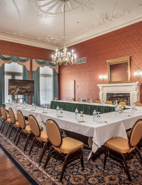 A formal conference room with a U-shaped table setup, cushioned chairs, ornate chandelier, red wall panels, and a decorative fireplace.