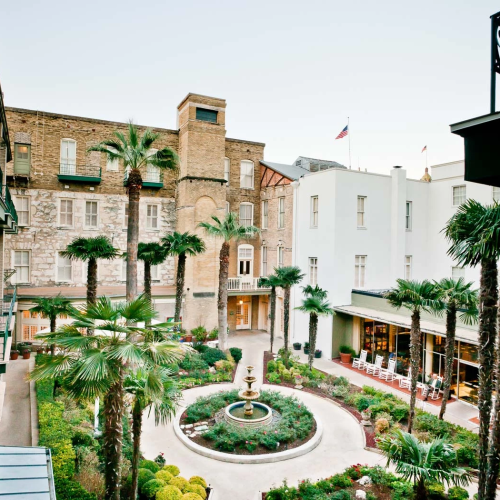 A sunny Mediterranean-style courtyard with lush potted plants, a stone walkway, and pastel buildings forming a cozy, intimate square.