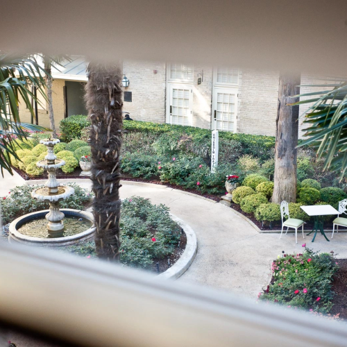 A balcony with wooden railings overlooks a tree-lined street; sunlight filters through leaves, and a parked car is visible below.
