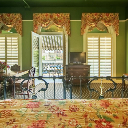 A vintage-style living room with green walls, floral curtains, a patterned bedspread/sofa set, and large windows letting in light.