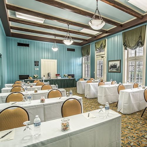 A bright, vintage-style dining room or small conference room with teal walls, long tables set with white linens, chairs, and place settings.