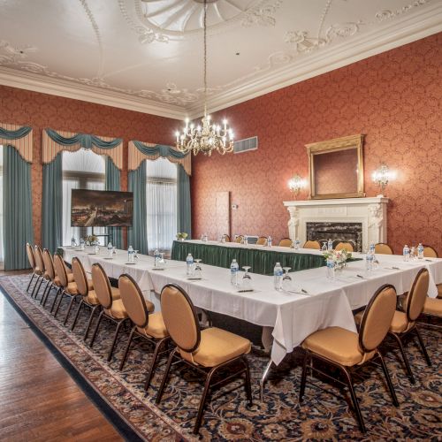 A formal conference dining room with a U-shaped table setup, green chairs, white tablecloths, chandeliers, and ornate red wallpaper.