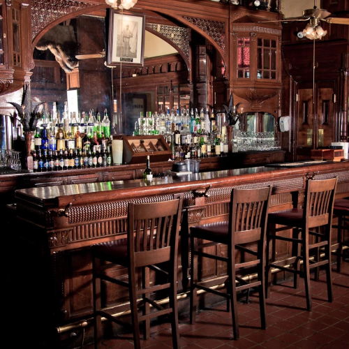 A classic wooden bar with a long counter, row of stools, shelves of bottles, and warm ambient lighting.