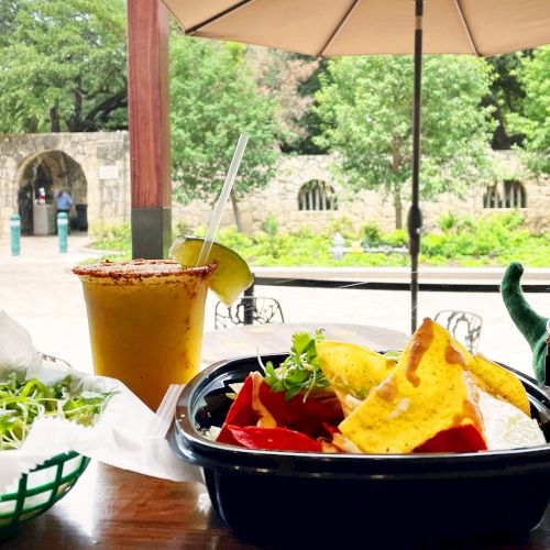 A plate of pickles and fresh greens, a bowl of curry, and a bottle of sauce on a colorful table outdoors, with a bowl of rice nearby.