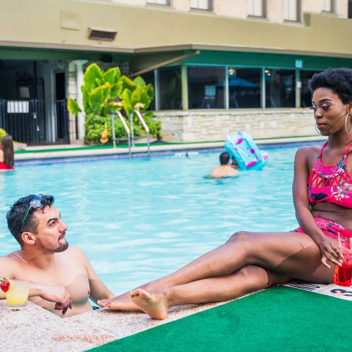 Two people relax at a pool edge: a man in swim trunks talks to a woman in a pink bikini seated on the deck.
