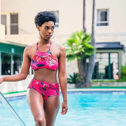A woman in a pink floral two-piece swimsuit stands by a pool, holding a rail and looking off to the side, sunny resort backdrop.