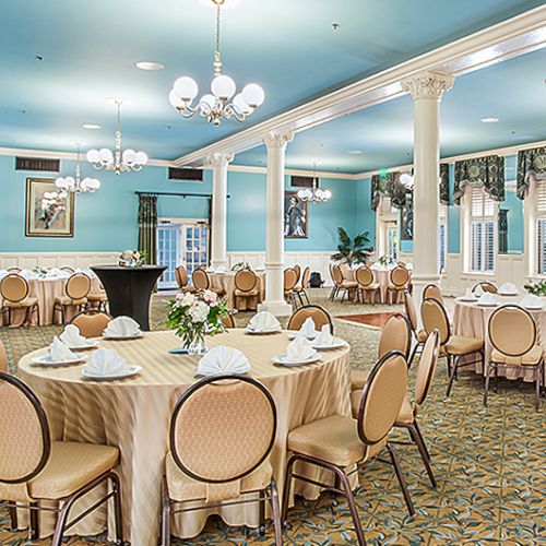 A bright, elegant dining hall with round tables, beige chairs, blue ceiling, chandeliers, and large windows; plants and decorative columns adorn the space.