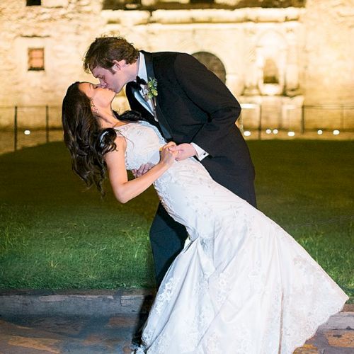 A newlywed couple sharing a kiss, the groom lifting the bride in a white gown as they smile, outside on a grassy area at night.