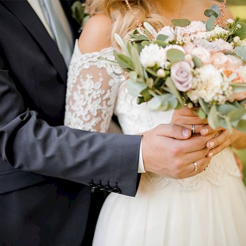 A bride and groom hold hands; the bride wears a lace wedding dress and holds a pastel bouquet, as the groom in a dark suit embraces her.
