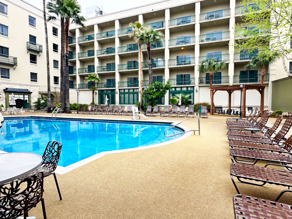 A resort-style hotel pool area with turquoise water, lounge chairs, a few tables, palm trees, and a multi-story beige building in the background.
