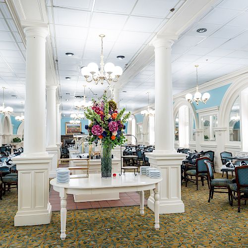 A bright, elegant dining hall with white columns, chandeliers, blue accents, and a central flower arrangement on a white table.