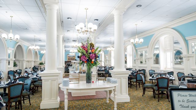 A bright, elegant dining hall with white columns, chandeliers, blue accents, and a central flower arrangement on a white table.