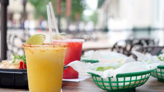 A sunny outdoor meal: a mango or orange smoothie with a spice rim, straw, lime wedge, plus bowls of food and chips on a communal table outdoors.